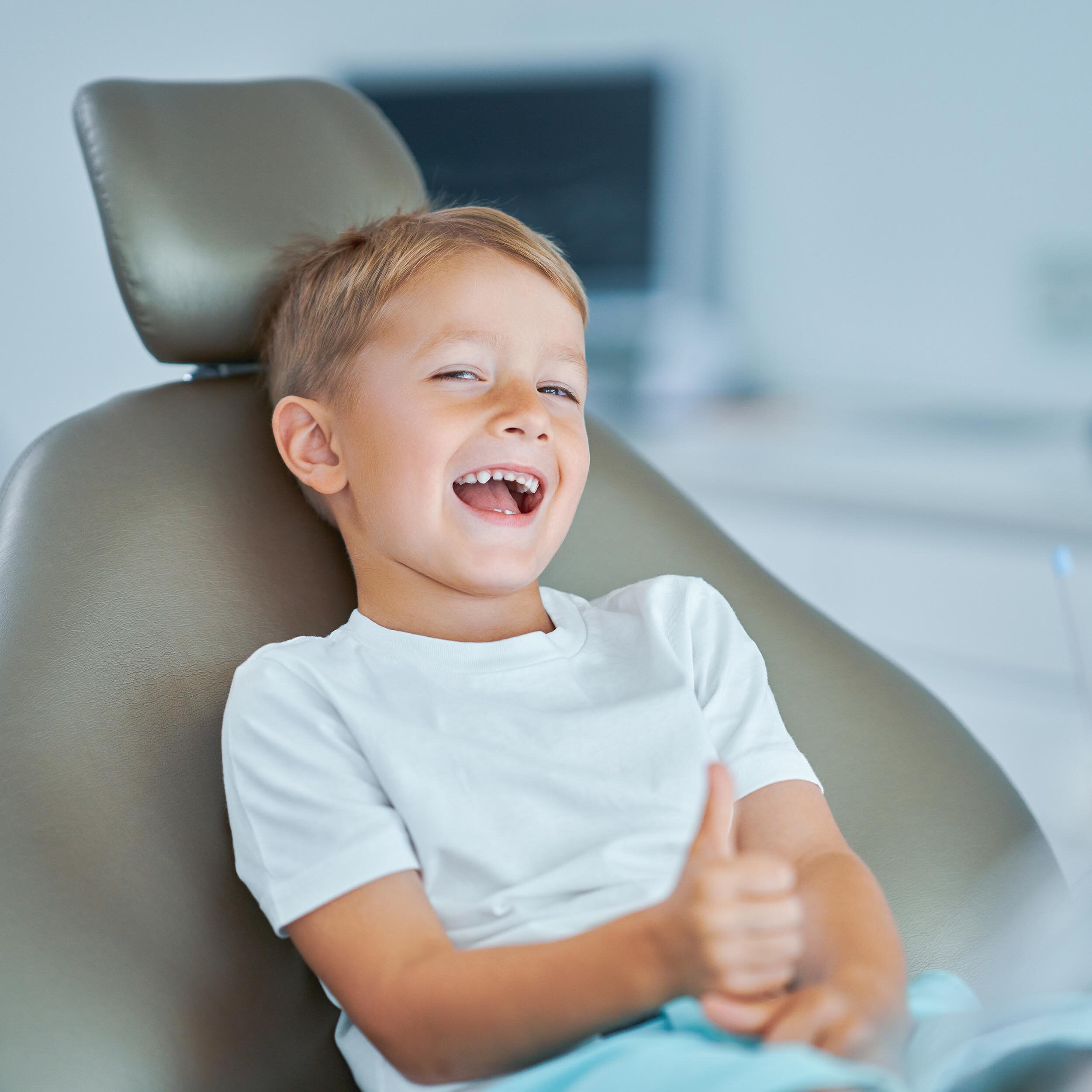 A young boy sitting in a dentist chair, giving a thumbs up.